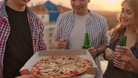 Young-russian-girls-and-boys-in-plaid-shirts-parses-pieces-of-hot-pizza-with-red-paper-and-eats-together.-This-is-a-rooftop-party-with-a-beer.